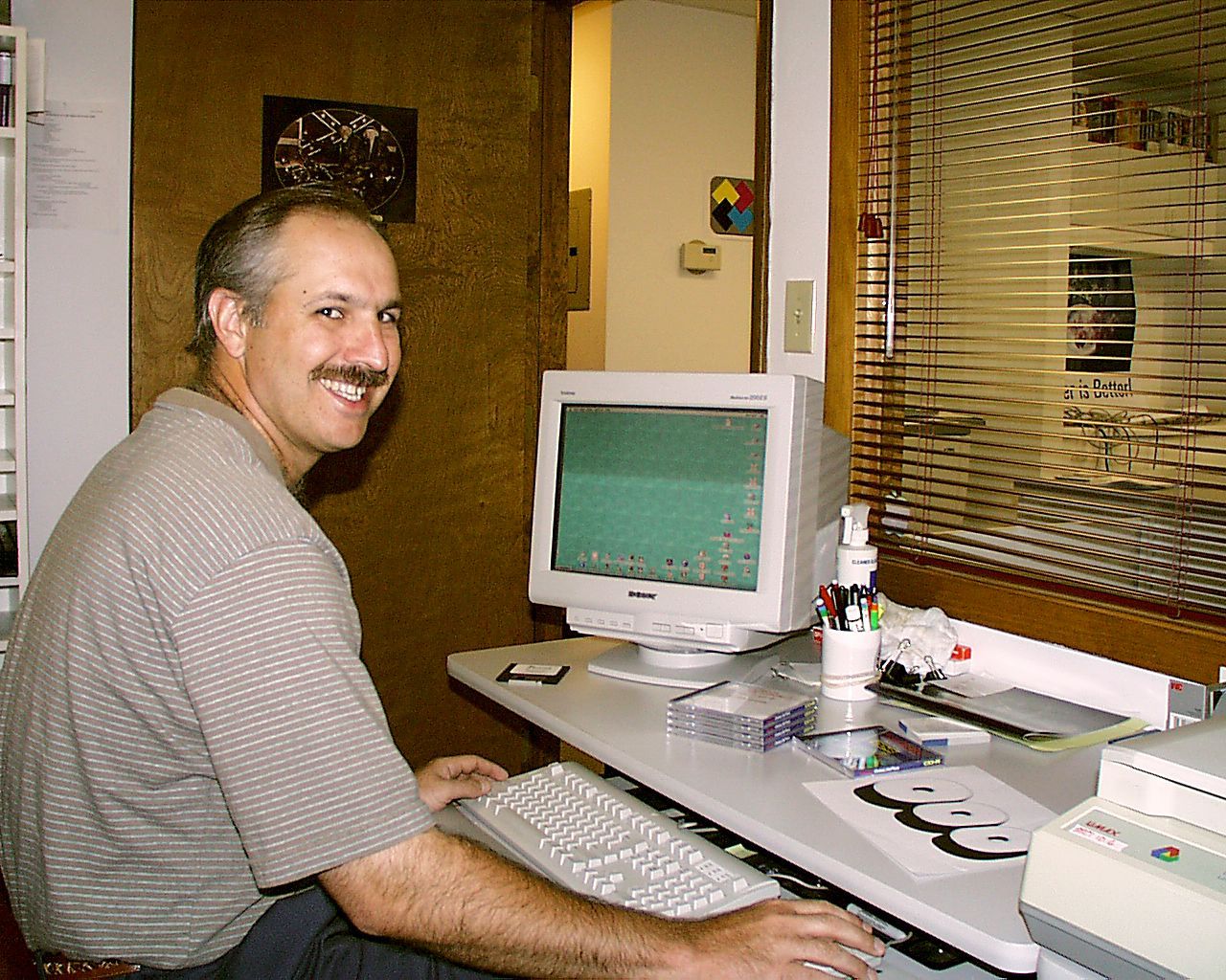 Man with a mustache smiles while using a computer at a desk in an office with a window and door.