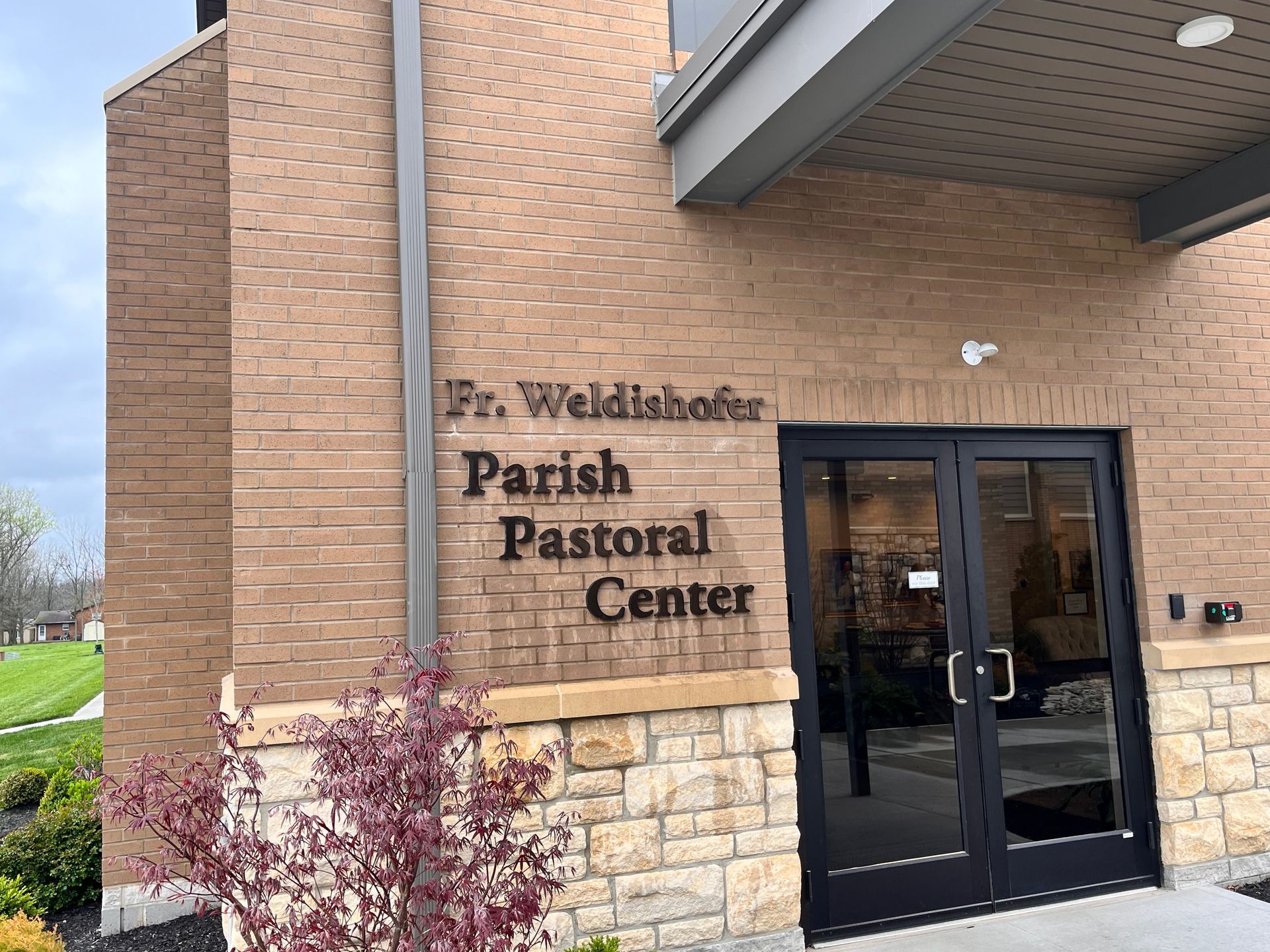 Fr. Wieldishofer Parish Pastoral Center entrance, brick building with black doors, tan stone accent, sign, and landscaping.