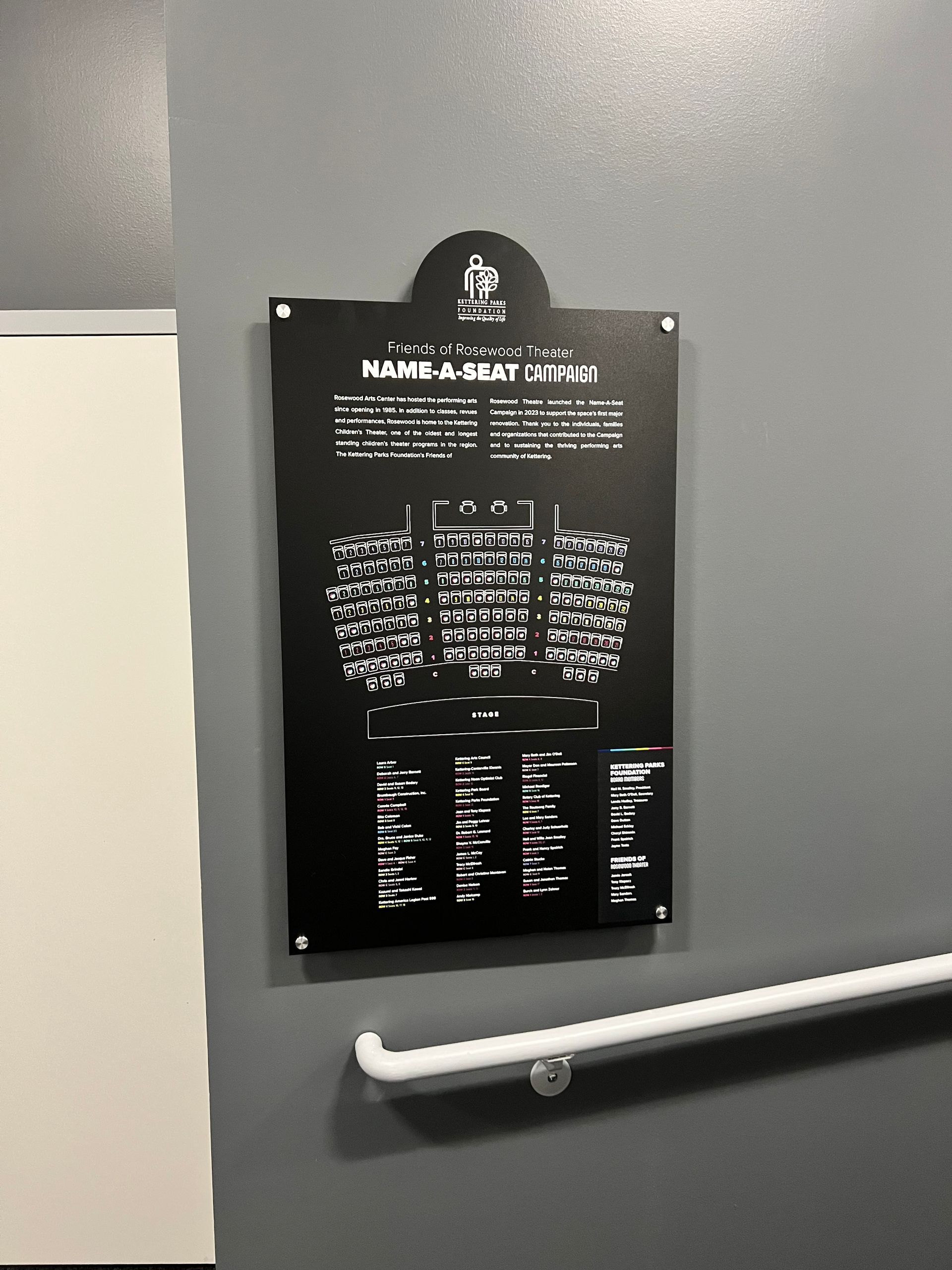 Black sign on a gray wall with white text detailing something, a white handrail is below it.