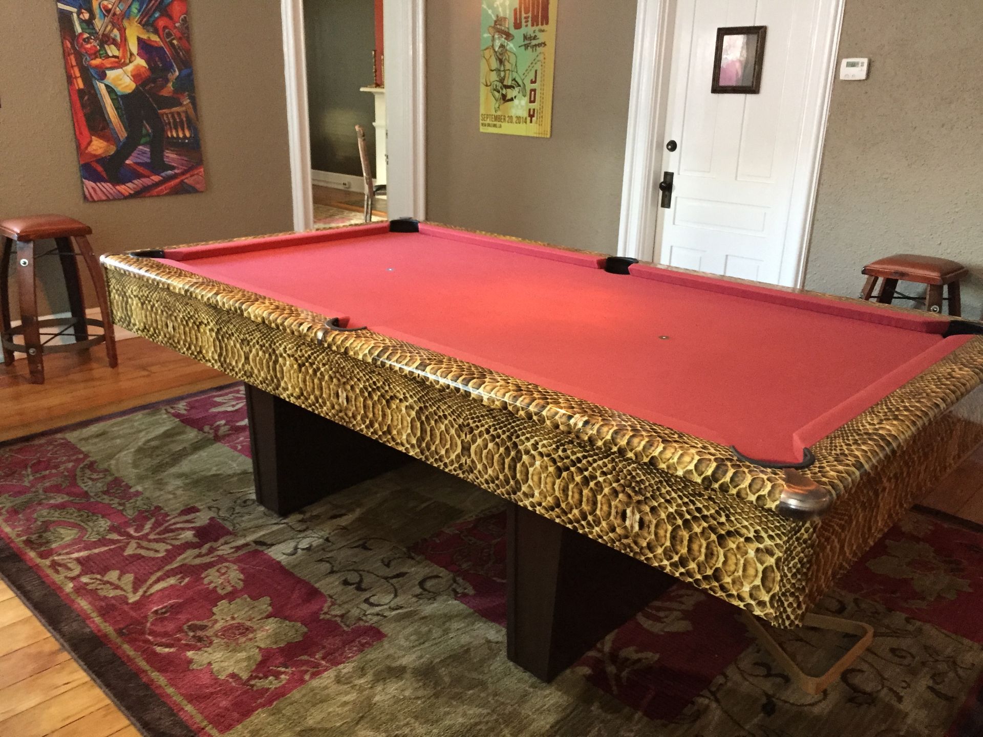 Pool table with red felt, snakeskin-like trim, and a vibrant patterned rug. Two stools and art are nearby.