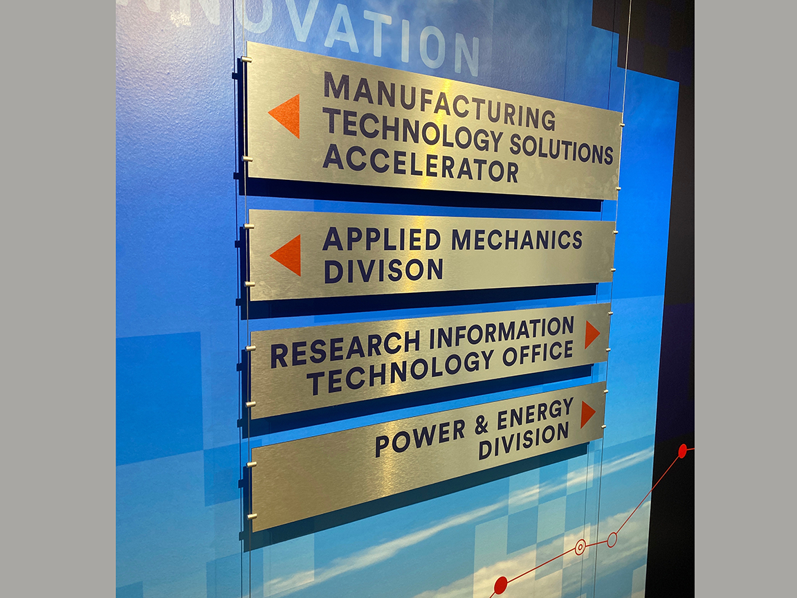 Sign with four metallic rectangular panels on a blue wall, listing departments: Manufacturing, Applied Mechanics, Research, and Power & Energy.