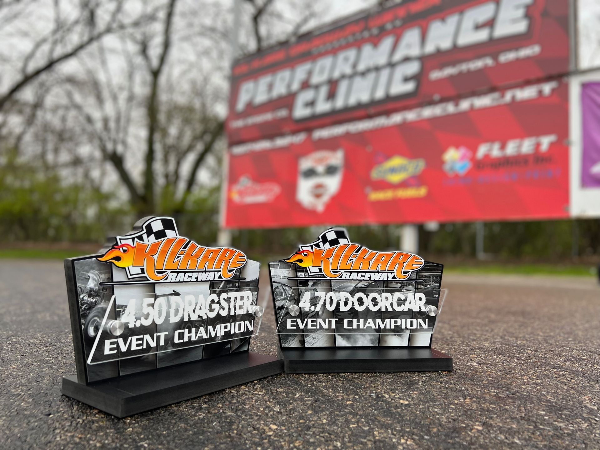 Two racing trophies: 4.50 Dragster and 4.70 Doorcar Event Champion. In front of a race track sign.