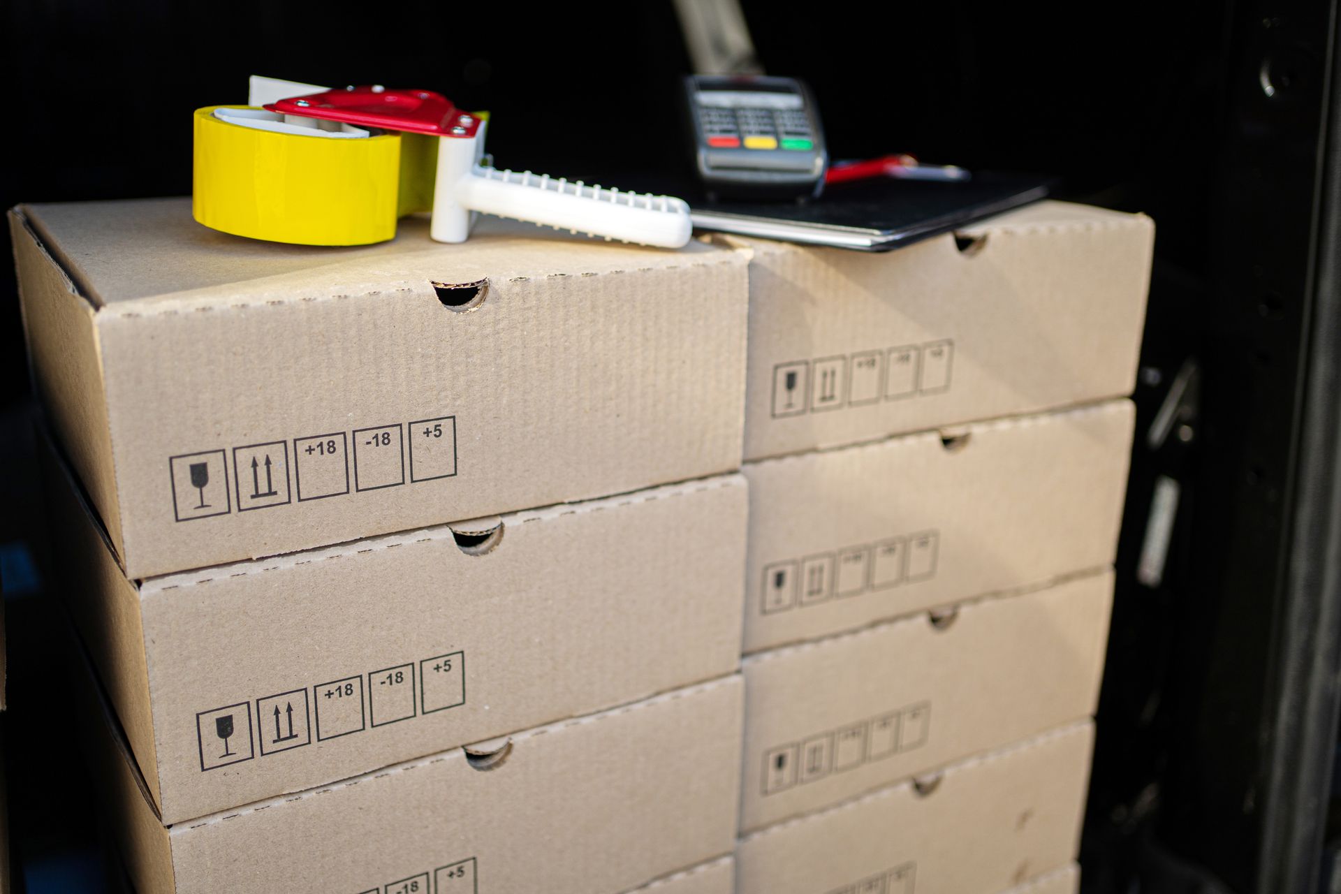 Stack of cardboard boxes with packing tape dispenser, yellow tape roll, and notepad.
