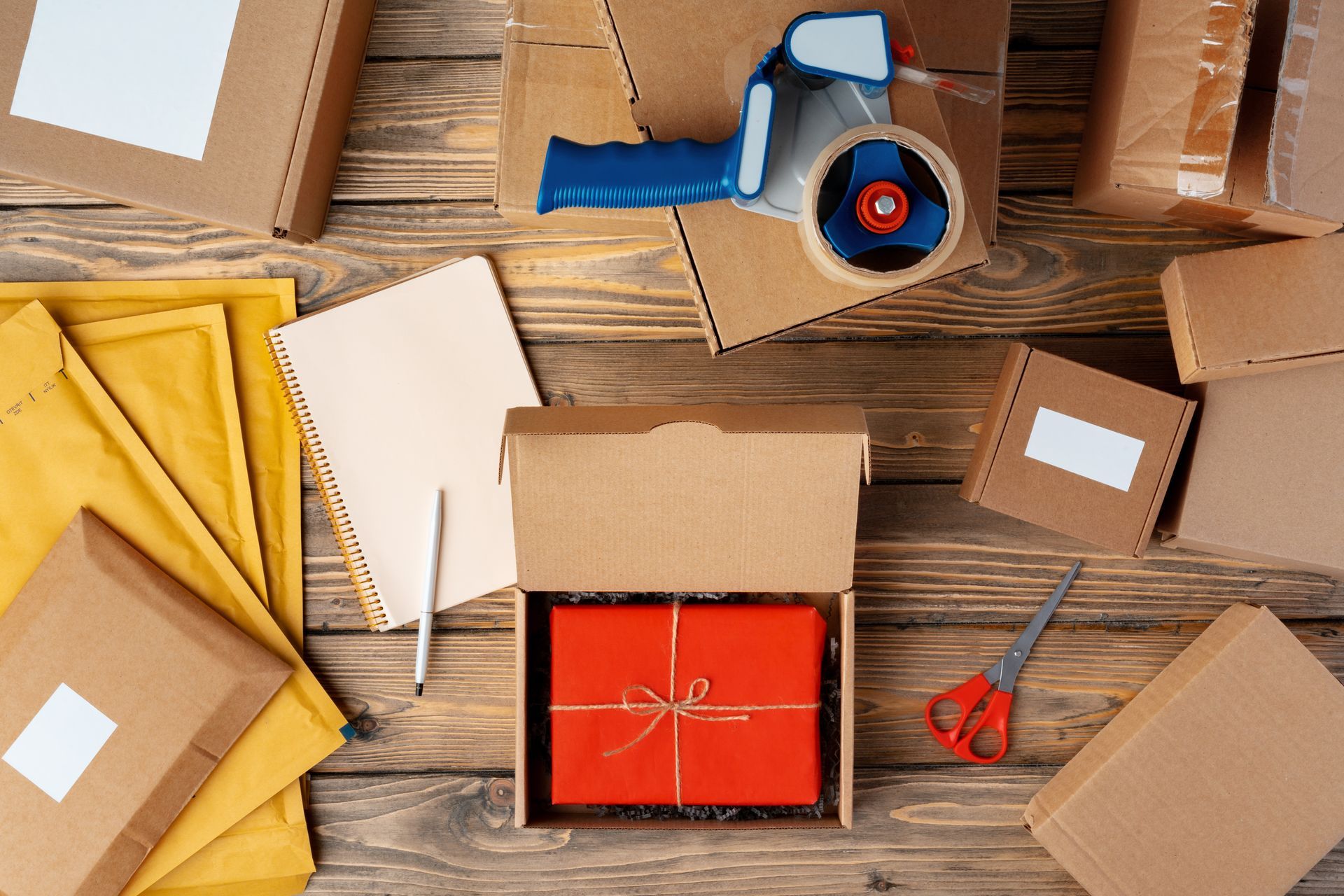 Various cardboard boxes, packing supplies, and a red gift inside a box on a wooden surface.