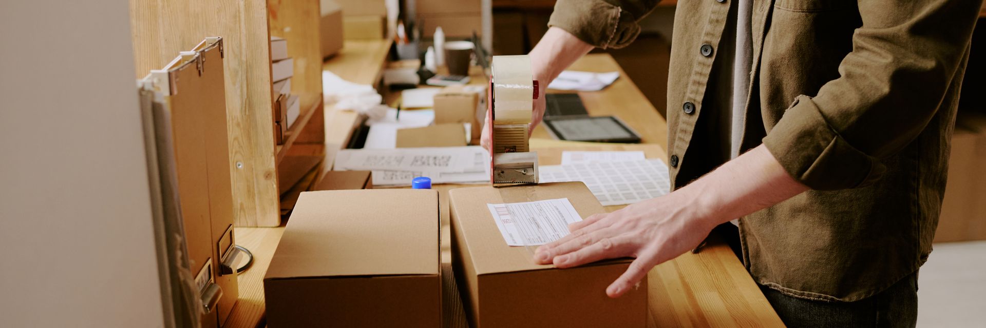 Person taping a cardboard box in a workspace. Several packages sit on a wooden table with a neutral background.