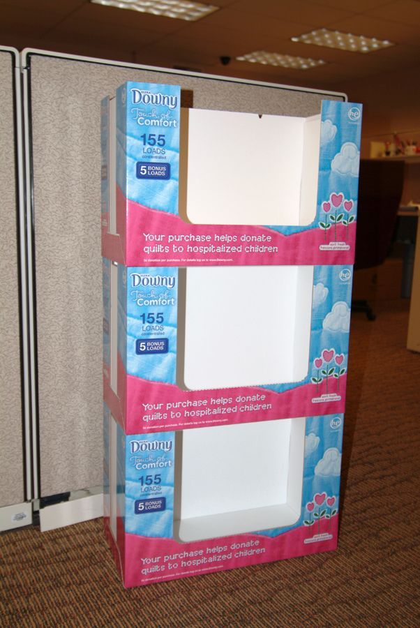 Three-tiered cardboard display for facial tissues on a carpeted floor, with blue and pink design.