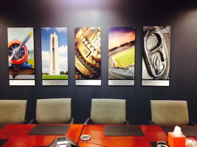 Five framed photos on a dark wall above a conference table. Images include plane, tower, gears, stadium, and can top.