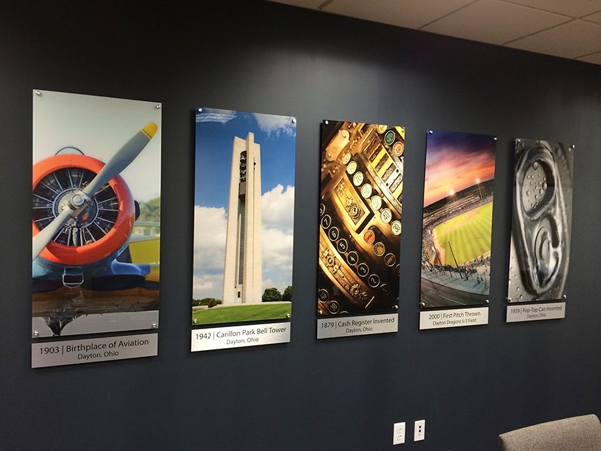 Five framed photos on a dark wall with titles below. Includes a plane, tower, cash register, stadium, and soda can.