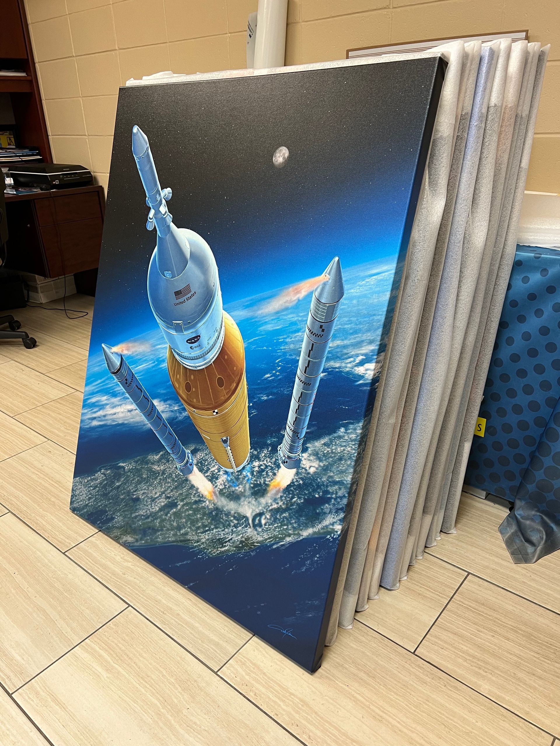 Artwork of a rocket launching from Earth, stacked with other framed art pieces.