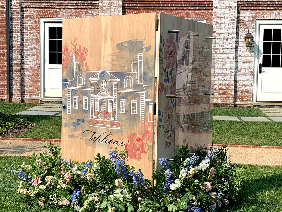 Wooden structure with painted house design, floral decor, in front of a brick building, on a lawn.