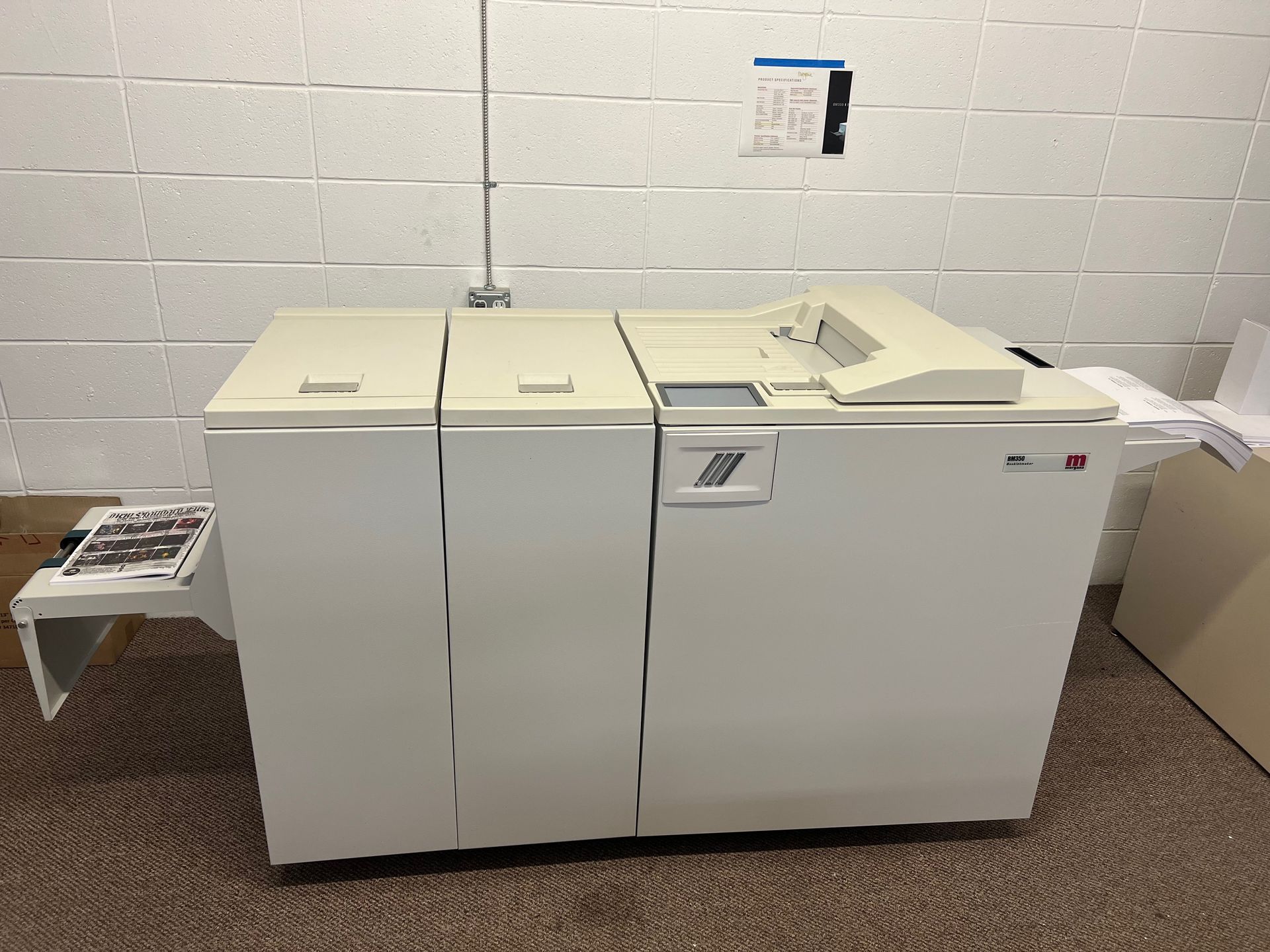 Large beige industrial printer against a beige brick wall; paper tray on side.