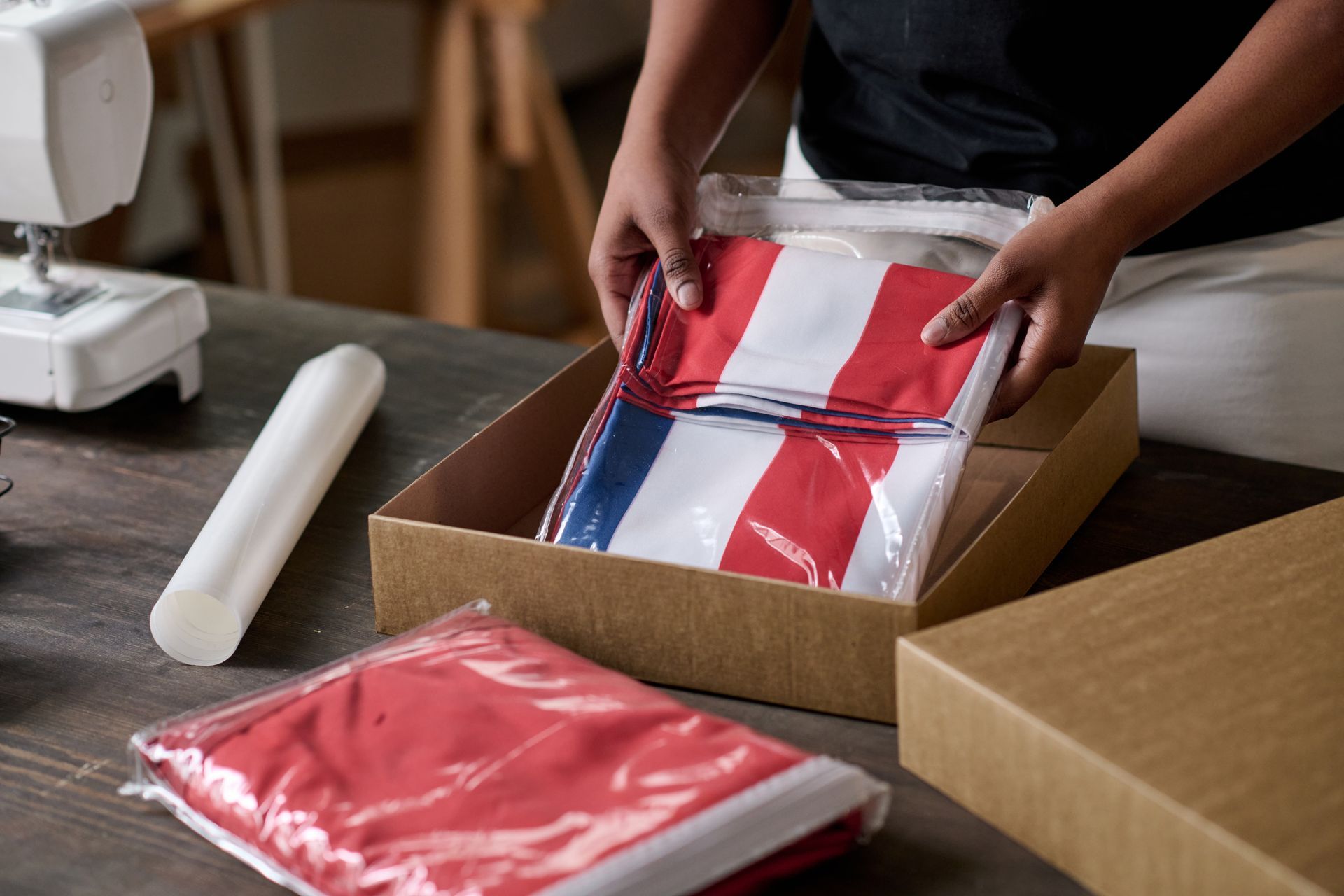 Person packing folded red, white, and blue striped flags in a box near a sewing machine.