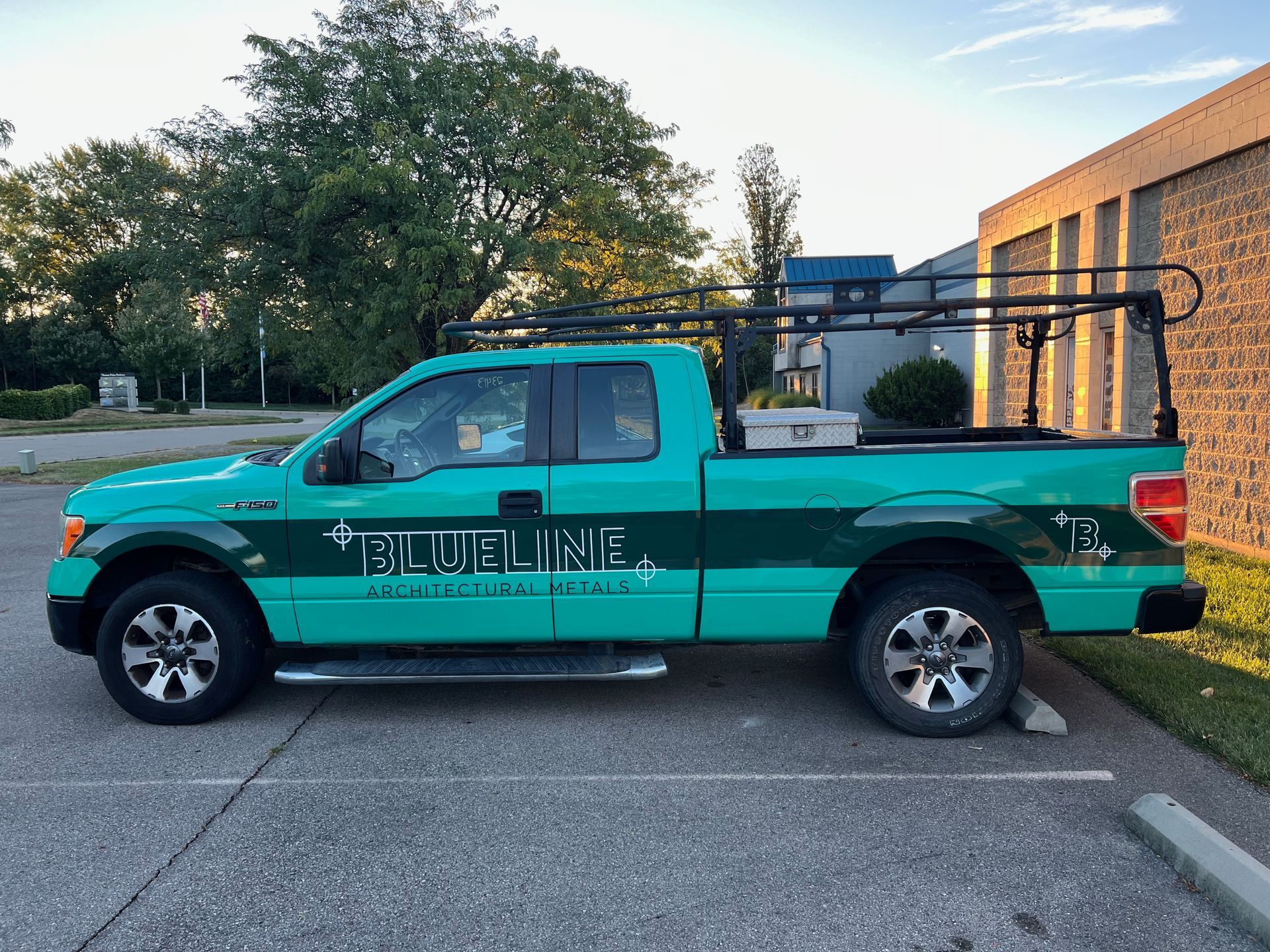 A teal Blue Line truck parked in a lot. It has a ladder rack and toolbox.