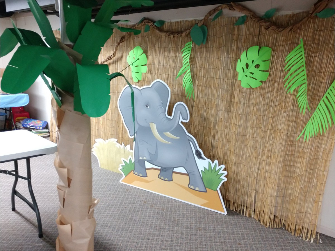 Jungle-themed decoration with a cardboard elephant cutout in front of a straw backdrop, palm tree on the left.
