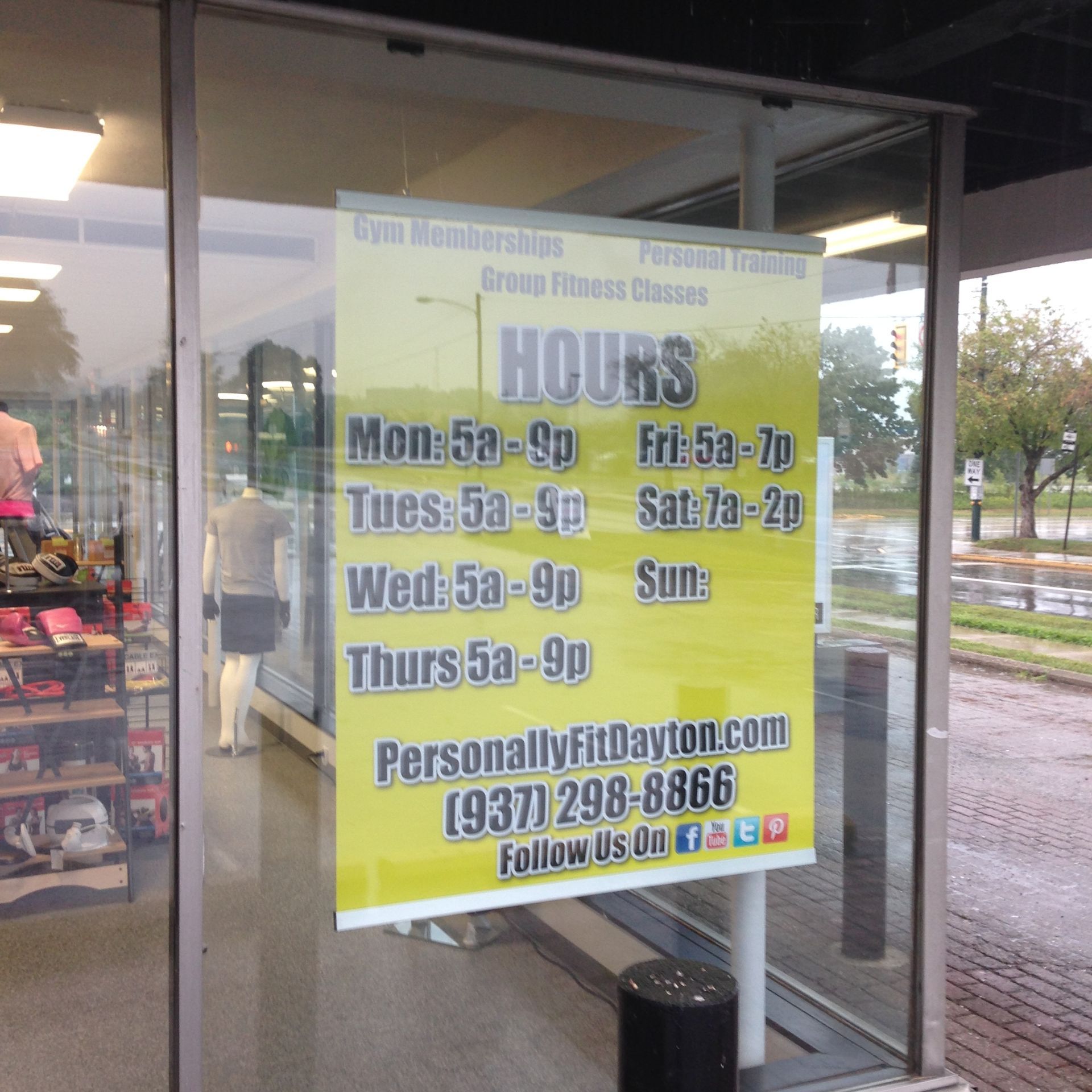 Sign on a fitness center's window listing gym hours and contact information; yellow background with black text.