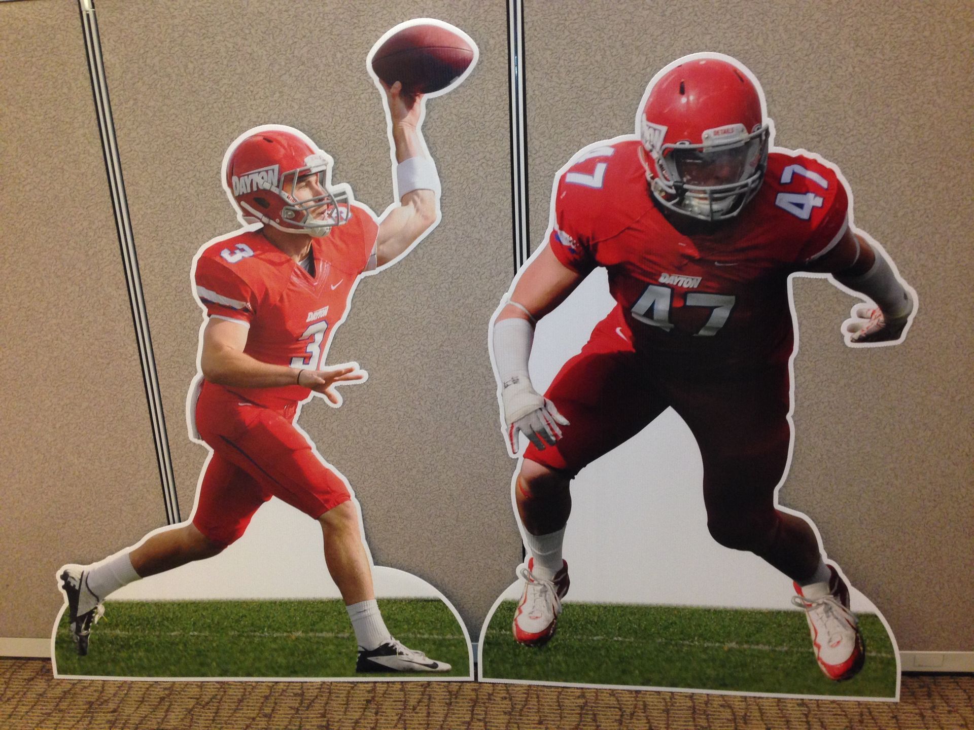 Two football players in red uniforms; one throwing, one in a defensive stance, both on a green field.