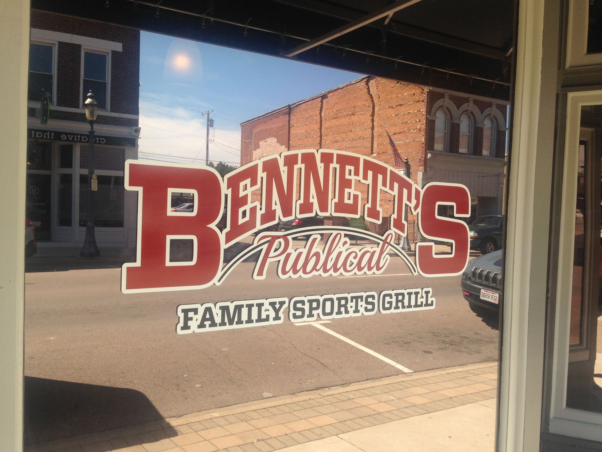 Bennett's Public House Family Sports Grill window sign with street view reflection, sunny day.
