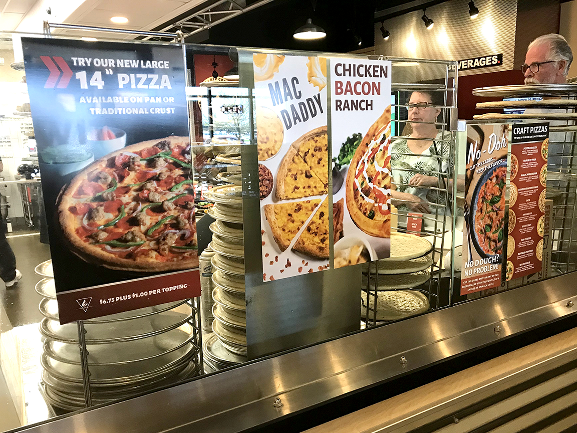 Pizza display with signs advertising different pizzas. A woman stands behind the counter.