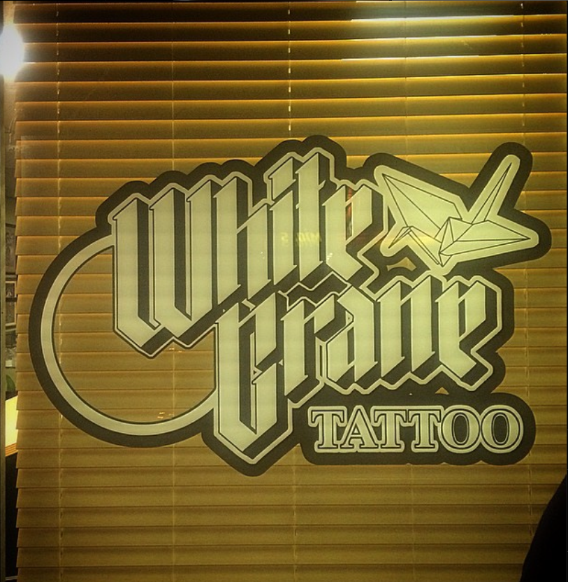 White Crane Tattoo sign with stylized lettering and origami crane on a window with blinds.