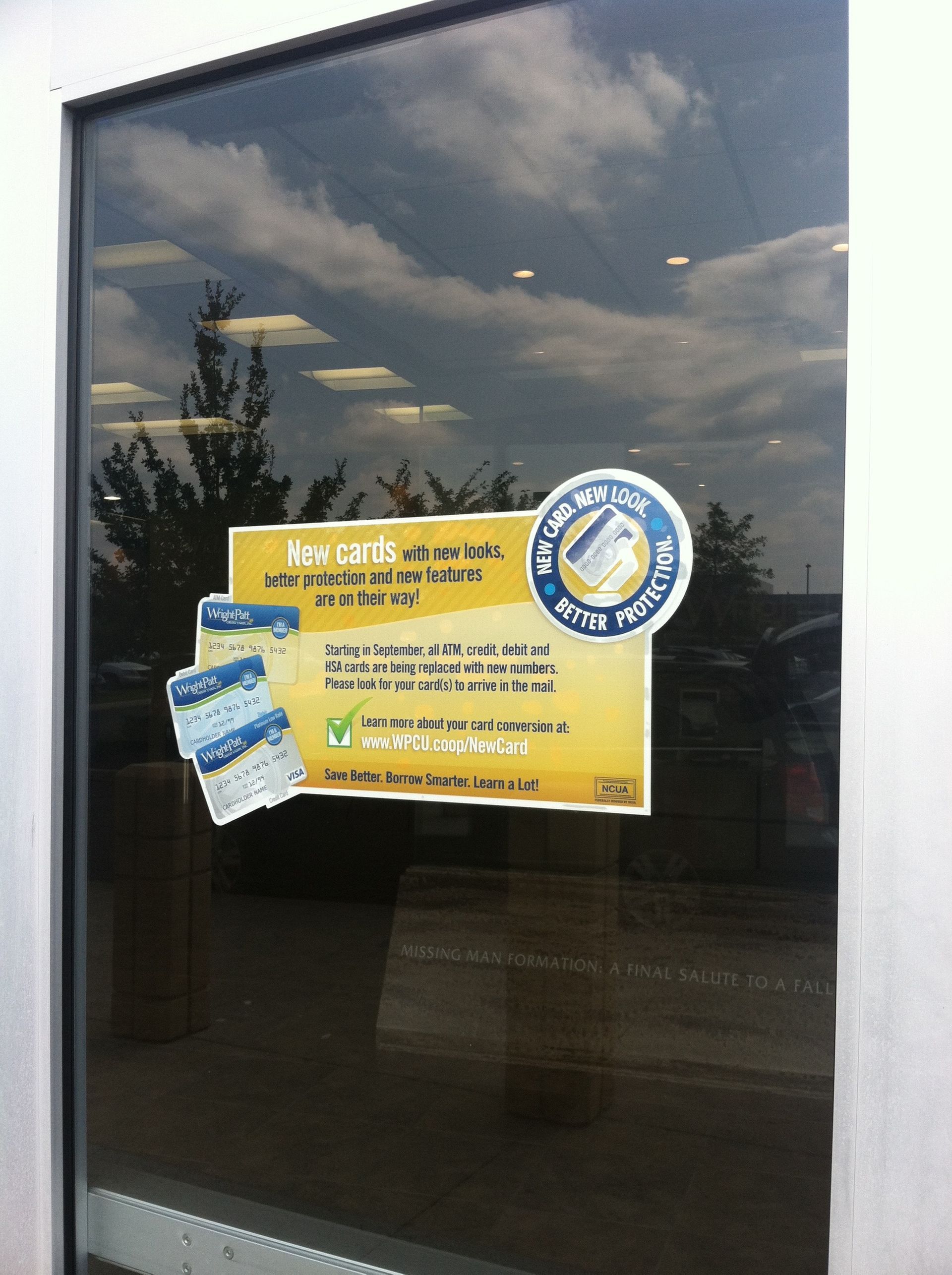 Door with a sign advertising card protection; a cloudy sky and trees are reflected in the glass.