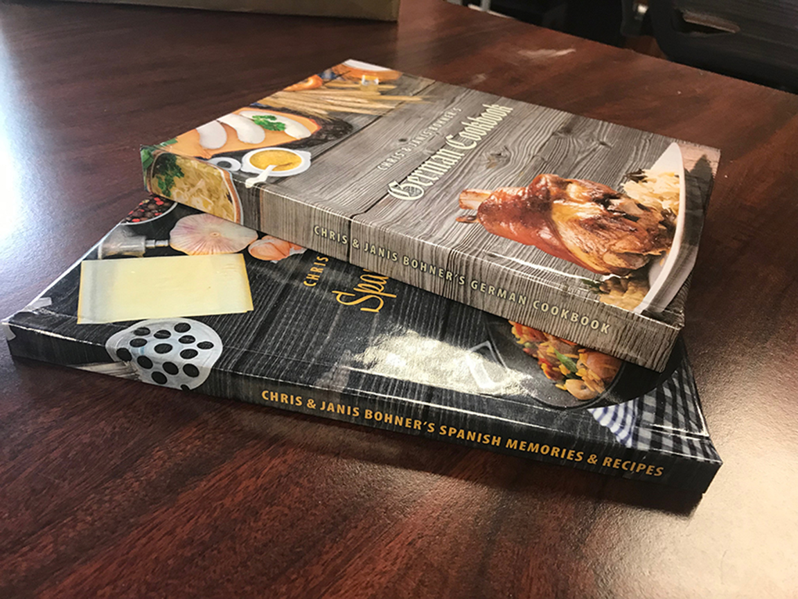 Two cookbooks stacked on a wooden table; one with sushi and cheese, the other with a ramen bowl.
