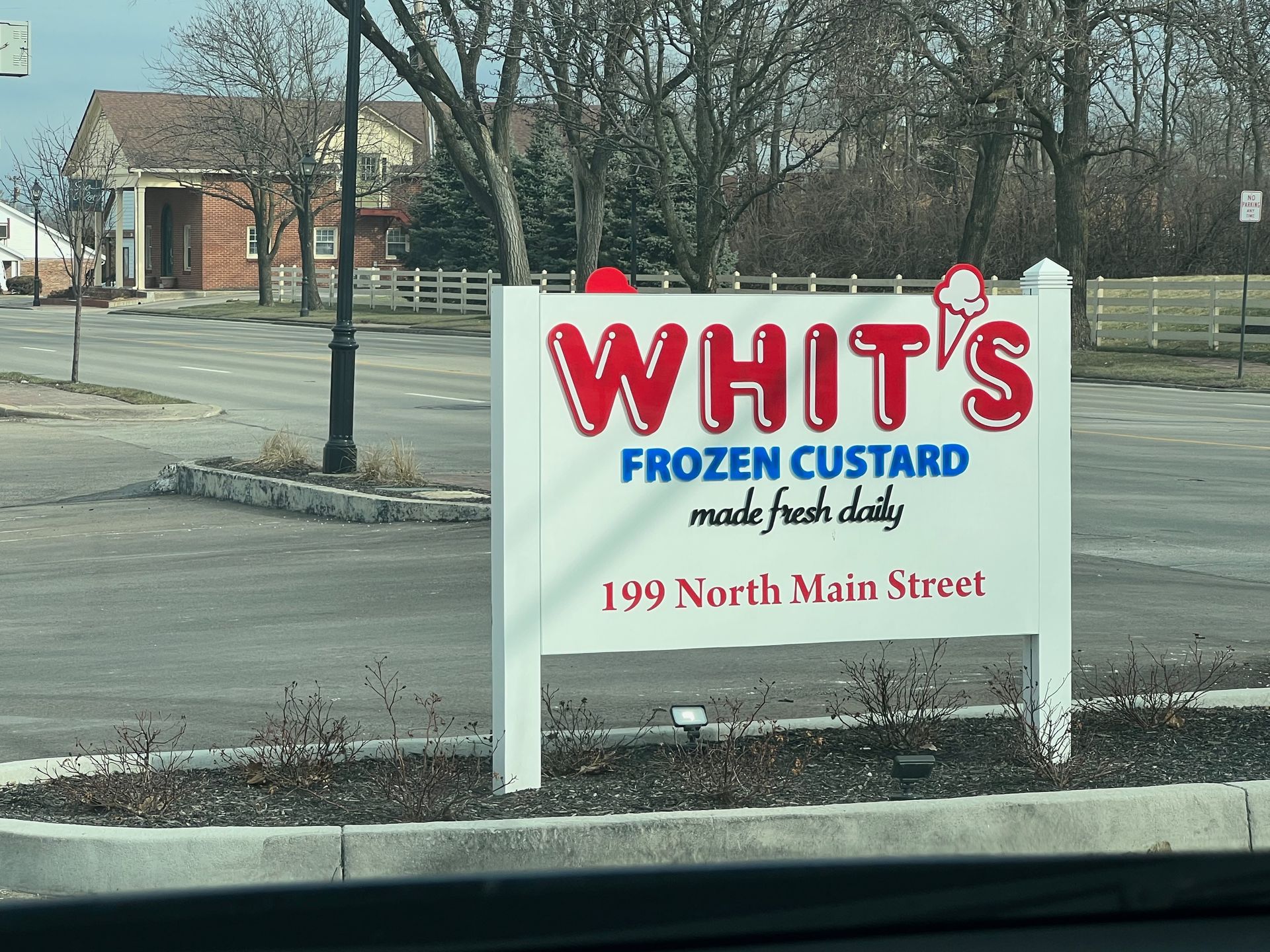 Sign for Whit's Frozen Custard, red and white, at 199 North Main Street, with building and trees in background.