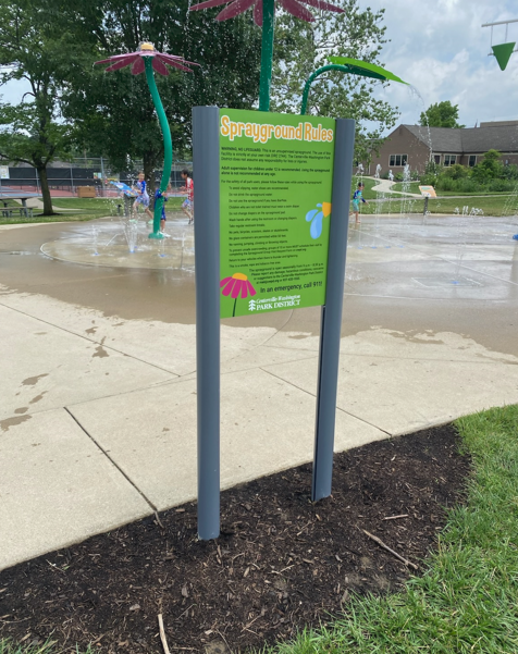 Sign with playground rules at a splash pad; green, pink and blue colors. People playing in the background.