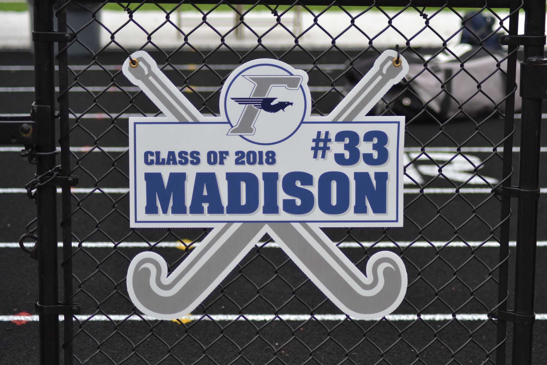 Sign for Madison, a 2018 graduate, with hockey sticks, school logo, and jersey number 33, displayed on a fence.