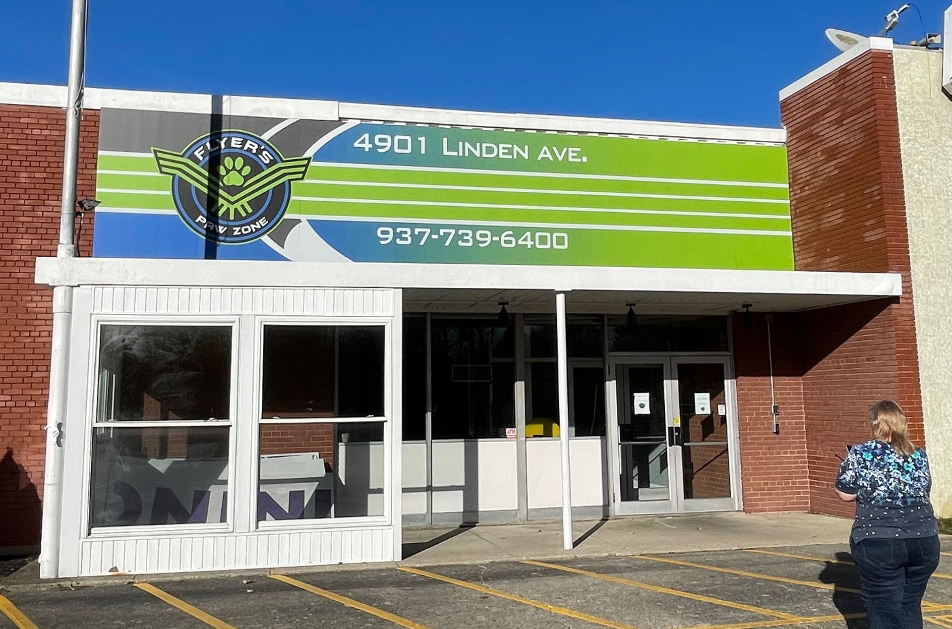 Exterior of a business, 4901 Linden Ave, with a green, blue, and black logo. A person stands to the right.