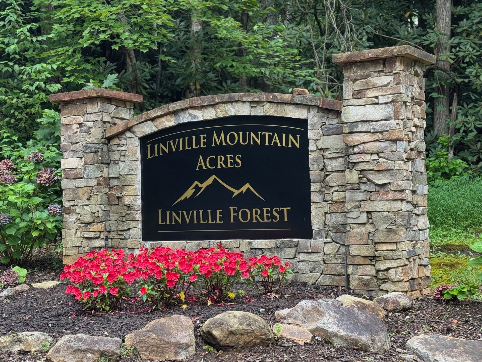 Stone entrance sign for Linville Mountain Acres/Linville Forest, with a black sign, gold text, and red flowers.