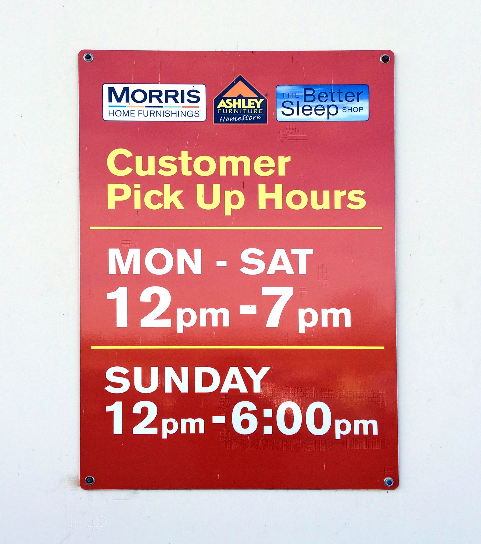 Sign indicating customer pick-up hours: Monday-Saturday 12pm-7pm, Sunday 12pm-6pm. Red background, white lettering.