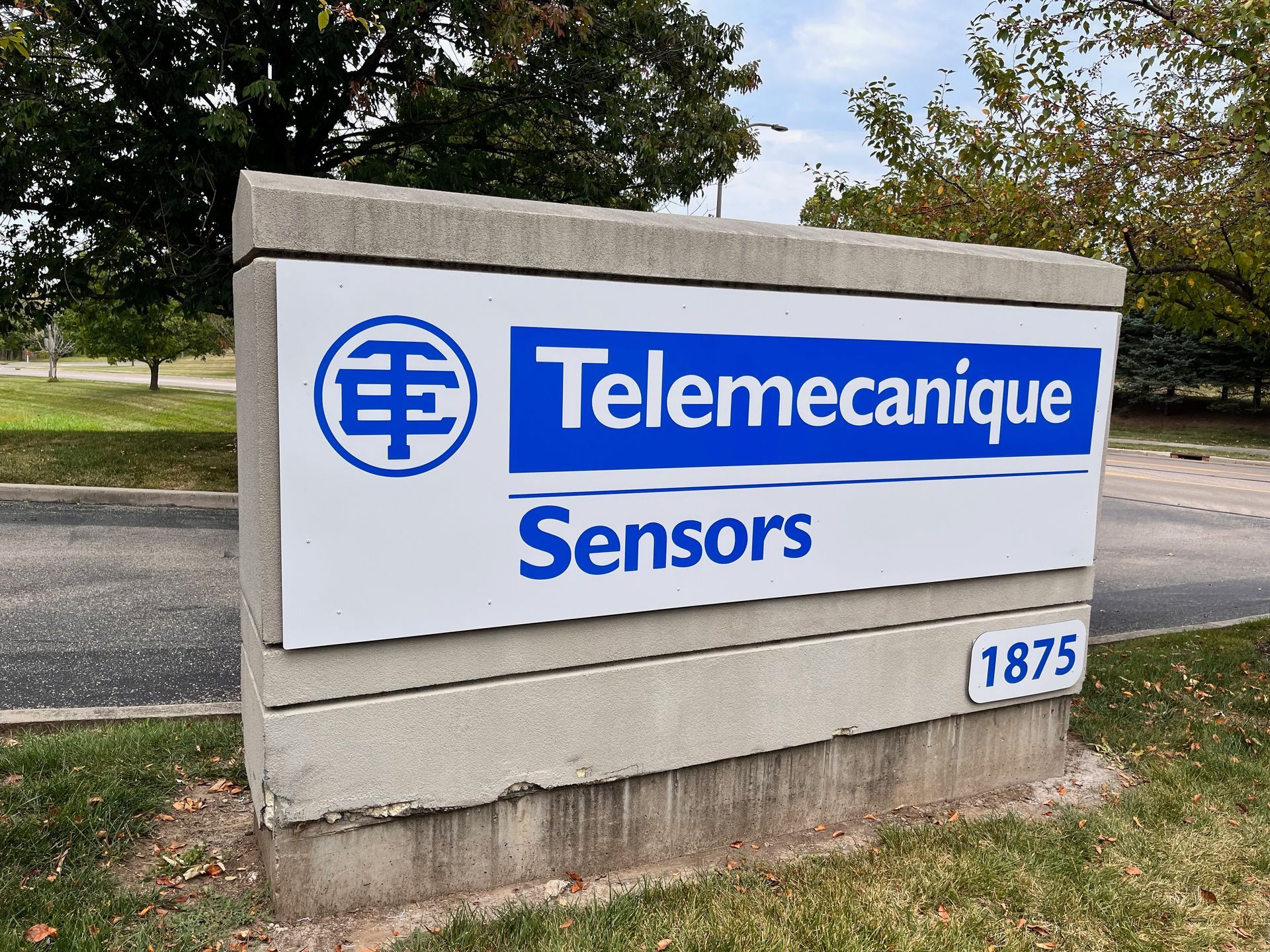 Sign for Telemecanique Sensors at 1875, blue and white on a concrete base, with green grass and trees.