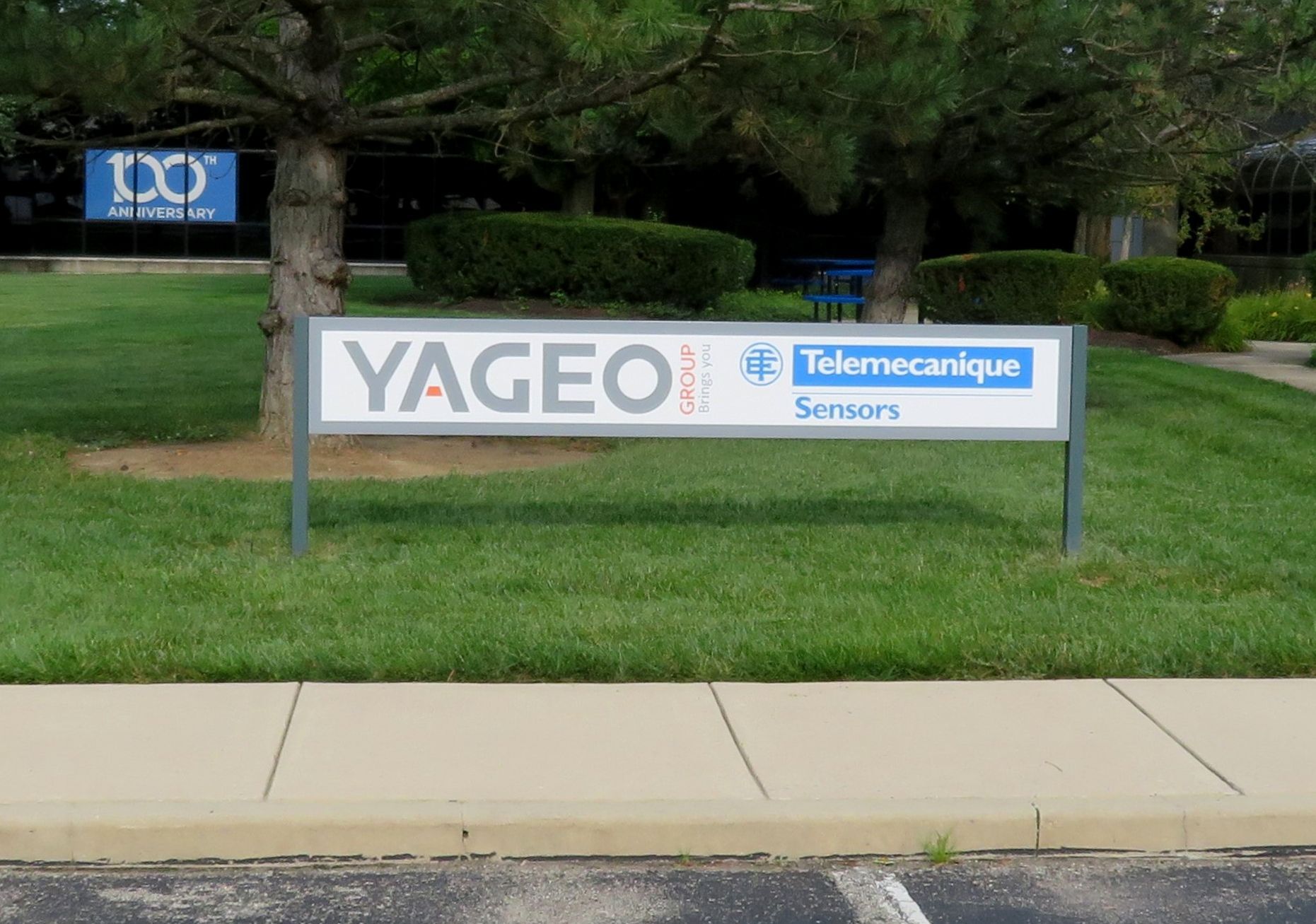 Sign for Yageo Sensors, with text in grey, red and blue, on green grass, next to a sidewalk.