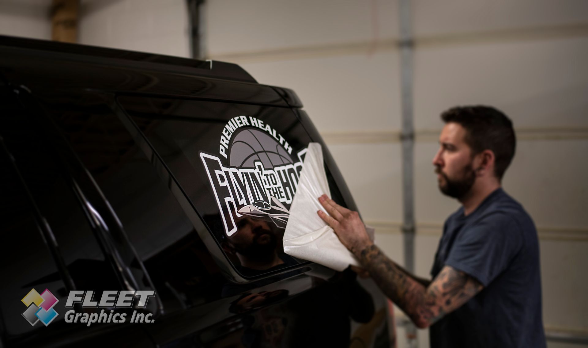 A man applying a white and gray decal on a black vehicle window in a garage.
