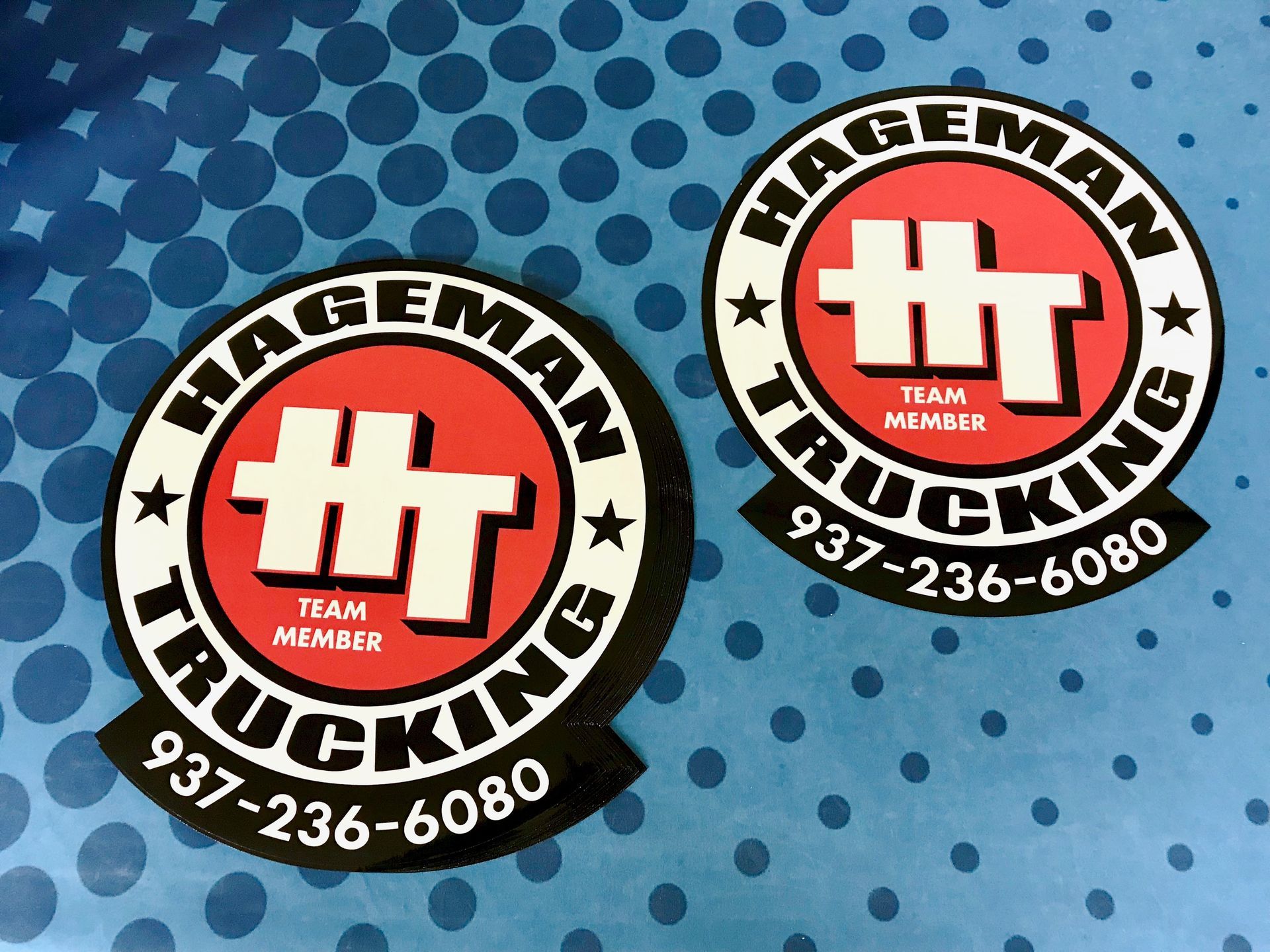 Two round Hageman Trucking stickers on a blue background with white dots. Each features a red center with 