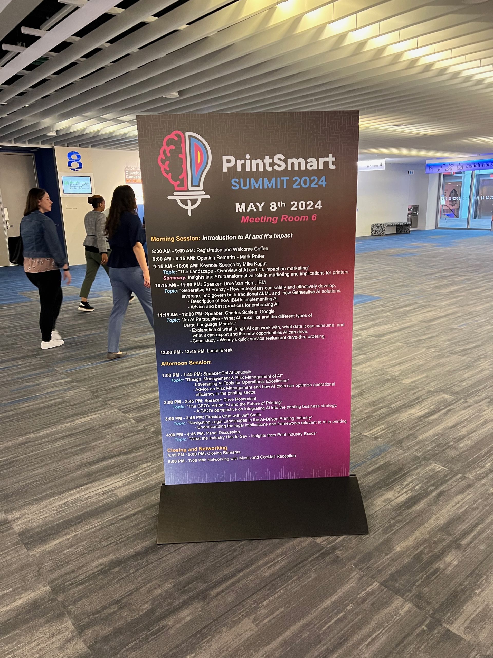 Sign for PrintSmart Summit 2024, May 2nd, with people walking by in an indoor setting.