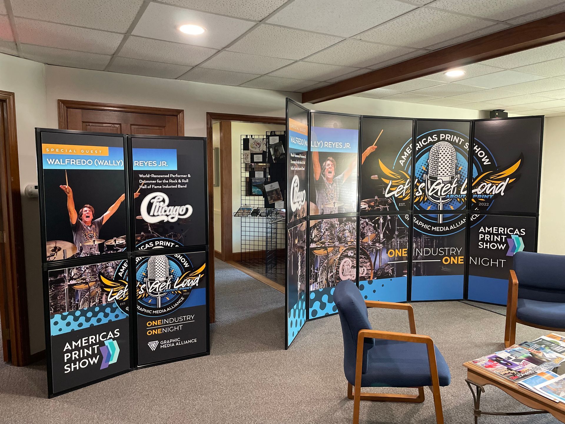A room displays promotional panels with event logos and photos. Blue chairs sit nearby.