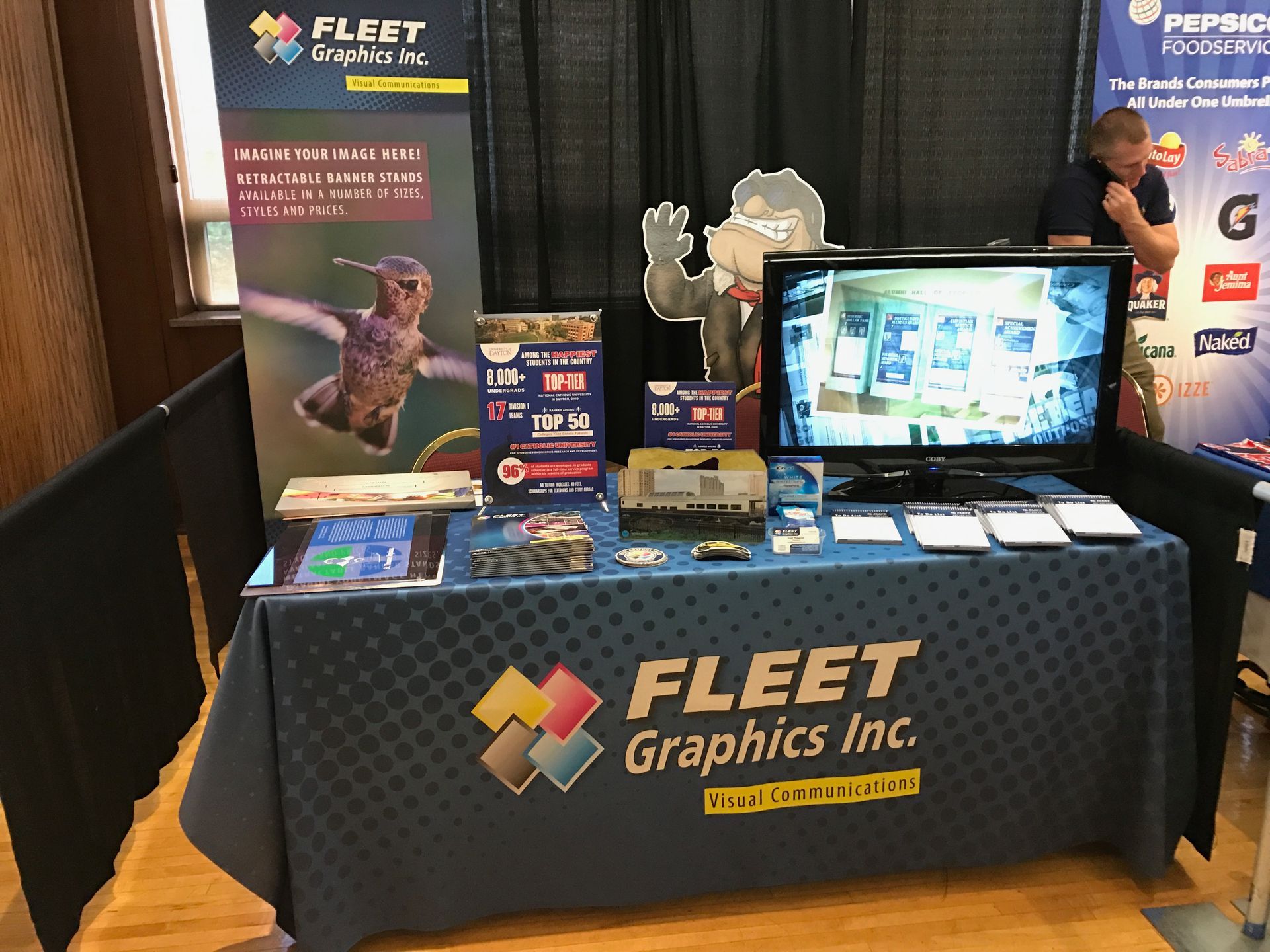 Trade show booth for Fleet Graphics Inc. Blue table with logo, display, and screen showing products.