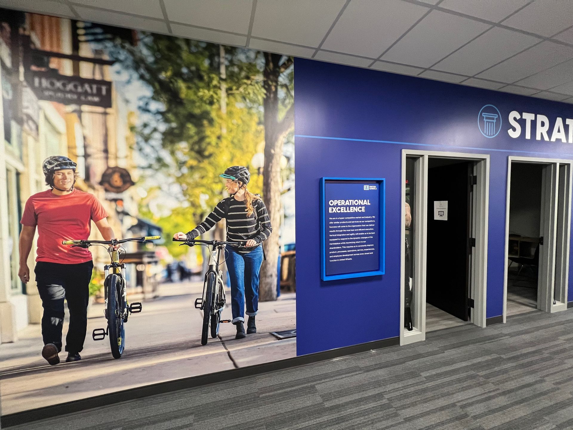 People with bikes walking, next to a blue wall with doors that say 'STRAT'.