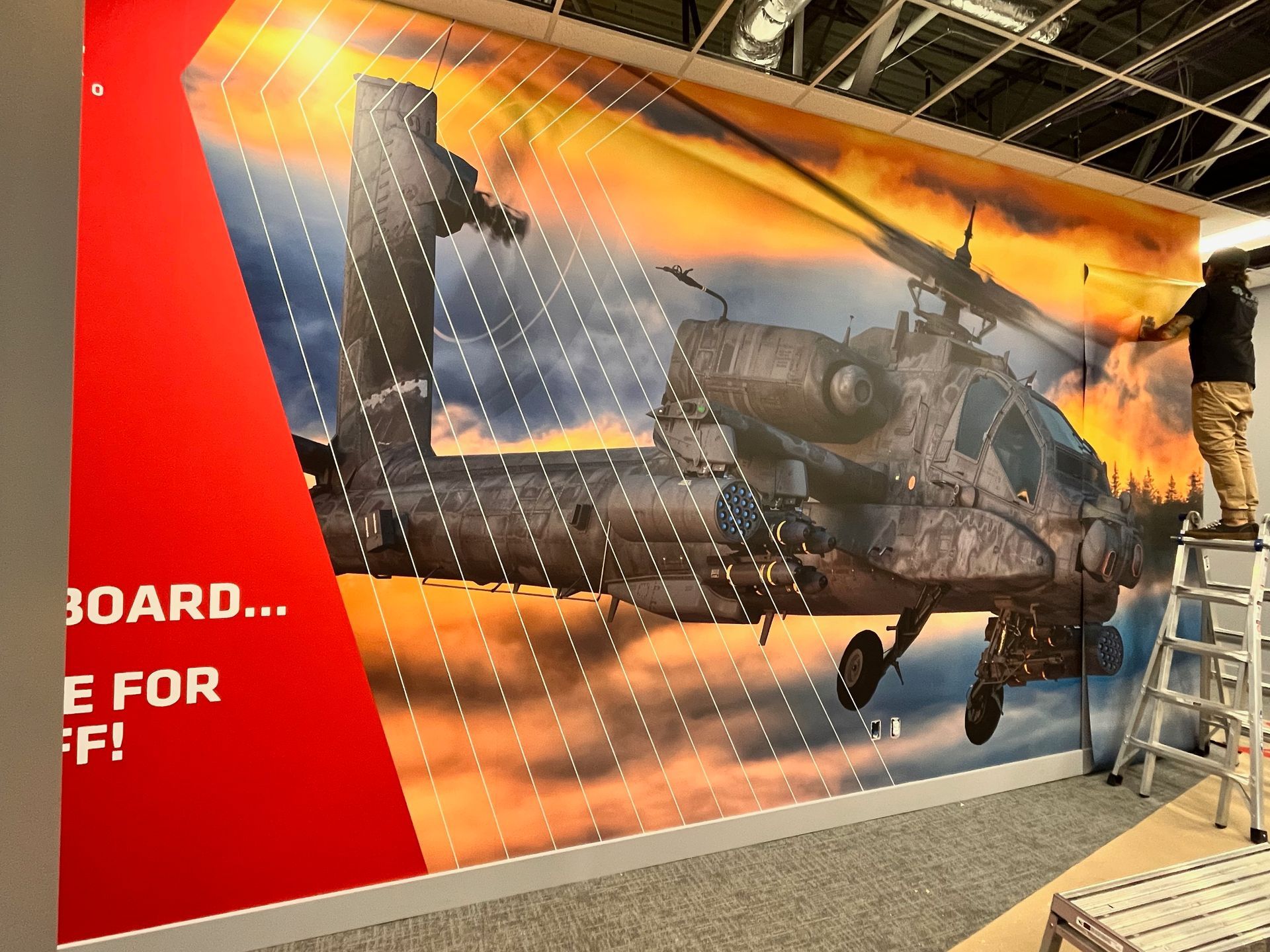Person on a ladder installing a large poster of a military helicopter. Bright orange, red, and white colors.