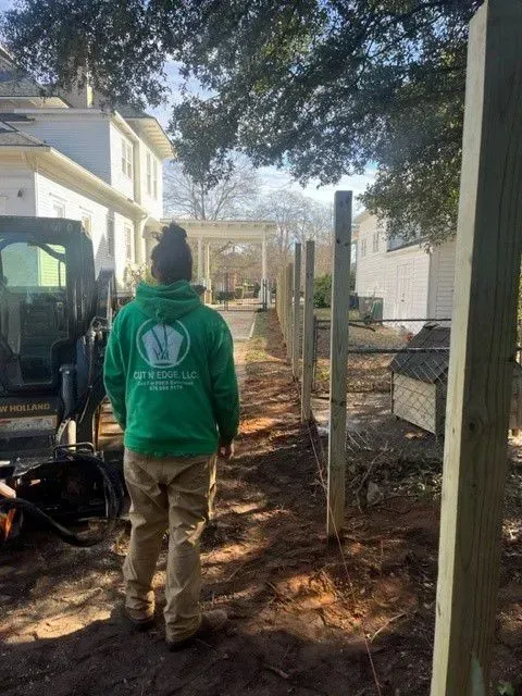 Person in green sweatshirt observing a partially constructed fence line, dirt path, and neighboring houses in the background.