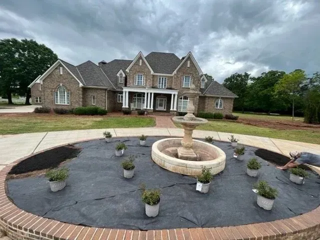Large brick house with fountain, surrounded by new plants on a cloudy day.