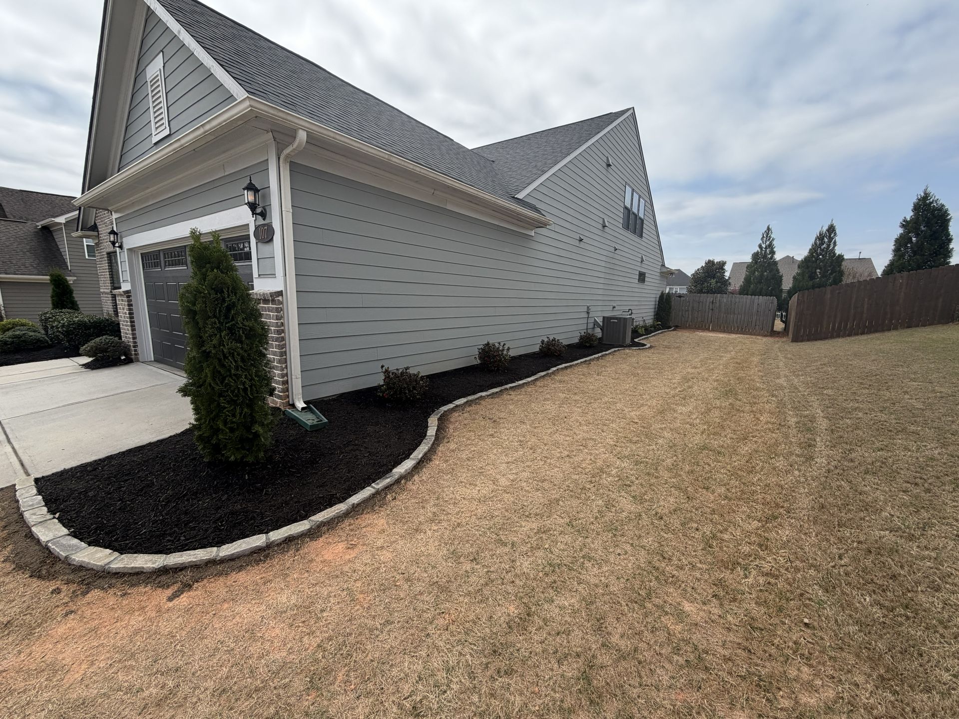 A gray-sided house with a garage, featuring a freshly mulched garden bed with stone edging along a dry, dormant lawn.