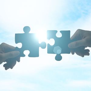 Two people are holding puzzle pieces in their hands against a blue sky.