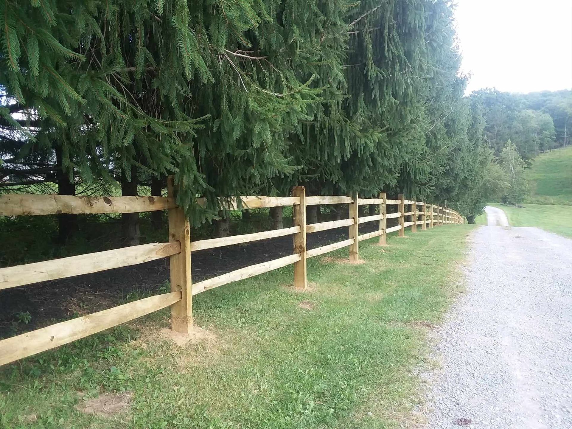 A wooden fence along a gravel road with trees in the background