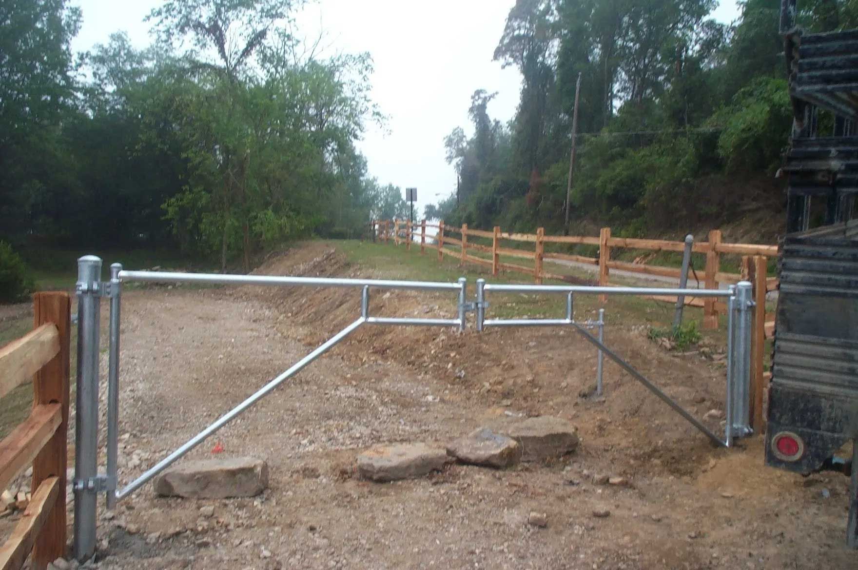 A metal gate is sitting in the middle of a dirt road next to a wooden fence.