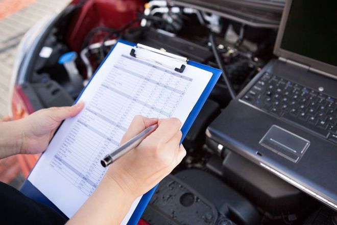 Hands filling out a checklist on a clipboard beside a car engine and laptop in a repair shop
