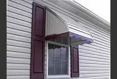 Window with burgundy shutters and a silver awning on a house with gray siding.