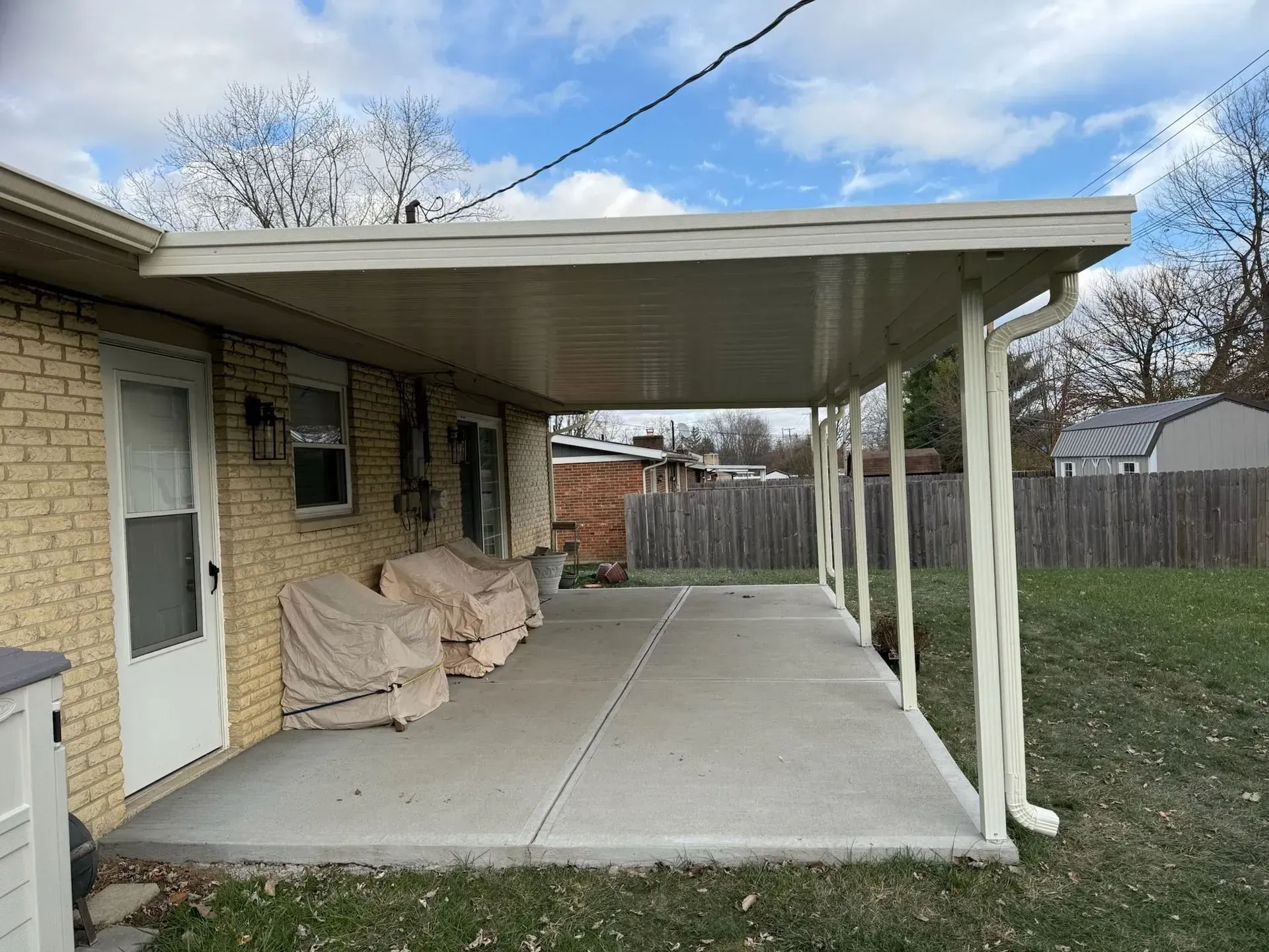 A covered patio attached to a brick house with concrete flooring, cream-colored roof, and support beams.