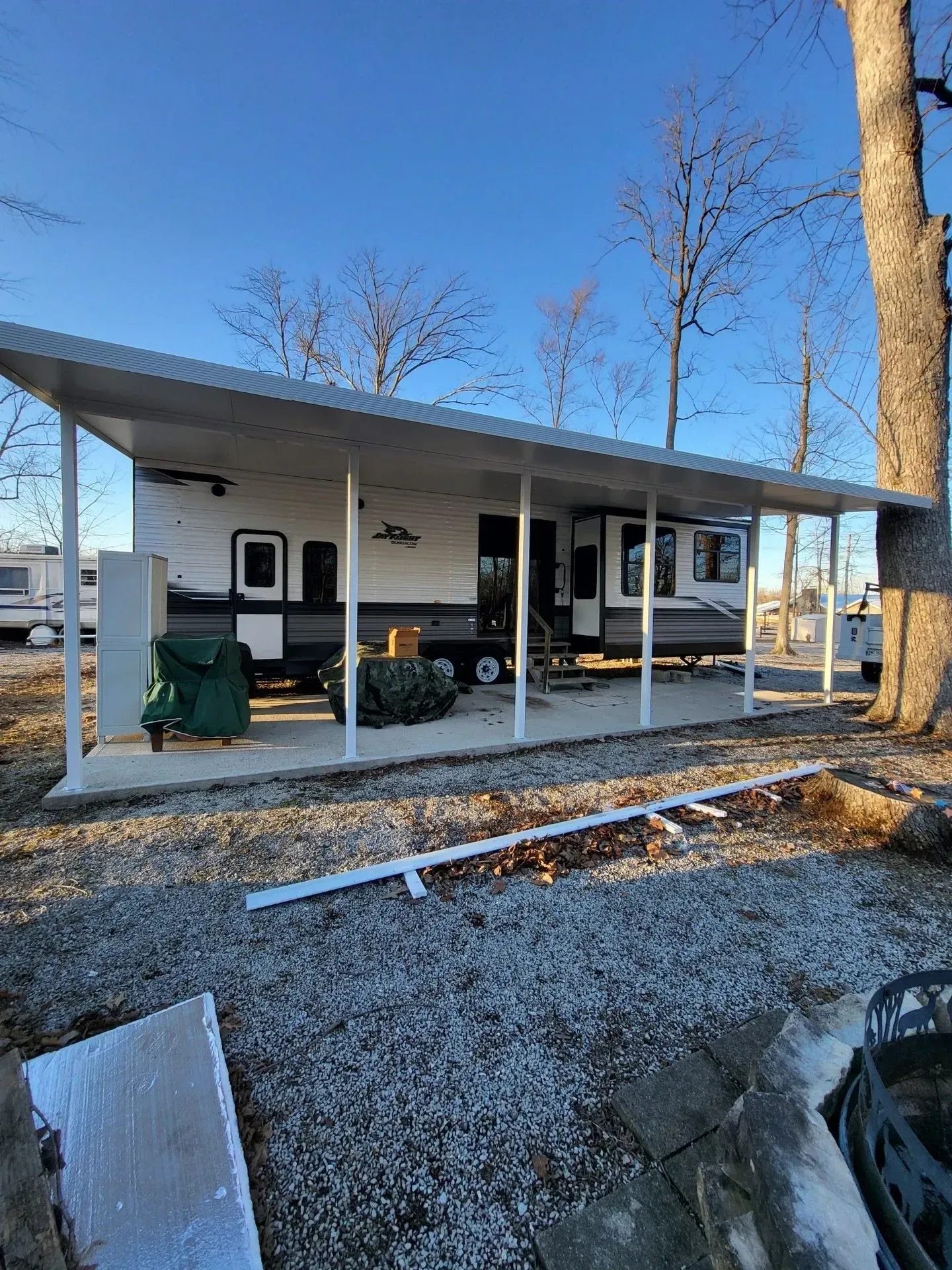 RV trailer under a white metal carport. Blue sky, bare trees, and a gravel ground.